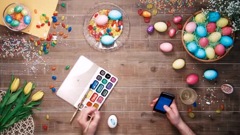 Man-painting-easter-egg-and-using-smartphone-on-table-decorated-with-easter-eggs.-Top-view