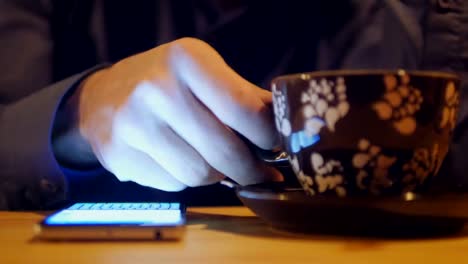 Close-up-of-man-hands-using-smartphone-for-messaging.-The-phone-rests-on-a-table-and-is-next-to-a-cup-of-coffee.-Man-drinks-coffee-and-chatting.