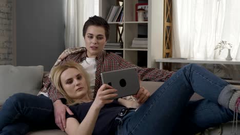 Lesbian-couple-is-resting-on-the-couch,-using-tablet-computer,-speaking,-showing-sign-no-by-shaking-head-60-fps