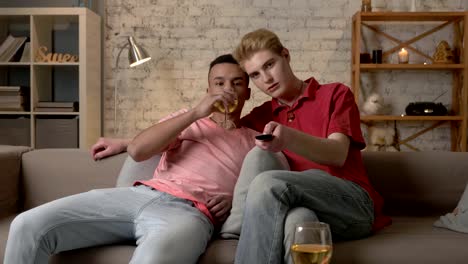 Multinational-gay-couple-sitting-on-couch,-watch-TV,-use-the-remote-control,-look-at-the-camera.-Homeliness,-romantic-evening,-cuddles,-hugs,-happy-LGBT-family-concept.-60-fps