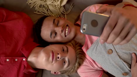 Happy-international-gay-couple-is-lying-on-the-couch-and-making-selfies-on-a-smartphone.-Homeliness,-LGBT-lovers,-happy-gay-family-concept,-laughing.-Say-hallo.-Video-chat.-Top-shoot-60-fps