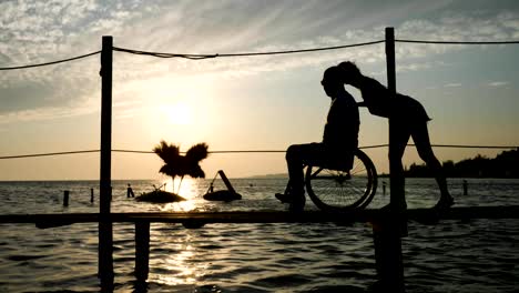 shapely-woman-with-Men-handicapped-on-wheel-chair-going-on-jetty-at-Embankment-against-heaven-in-afterglow