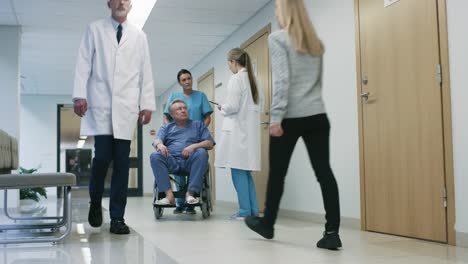 In-the-Hospital-Hallway,-Nurse-Pushes-Elderly-Patient-in-the-Wheelchair,-Doctor-Talks-to-Them-while-Using-Tablet-Computer.-Clean,-New-Hospital-with-Professional-Medical-Personnel.