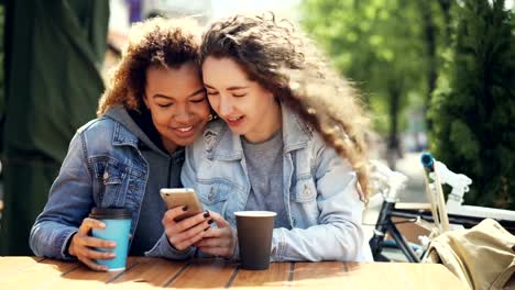 Attractive-young-ladies-Caucasian-and-African-American-are-using-smartphone-looking-at-screen-and-laughing-sitting-in-outdoor-cafe-at-table-together.