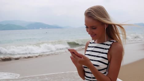 Young-beautiful-slim-woman-with-long-blonde-hair-in-black-and-white-dress-standing-on-the-coast-and-using-smartphone-over-background-at-storm-on-the-sea..-Girl-on-the-beach-touching-screen-and-smile.