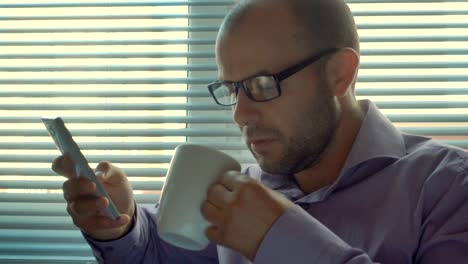 Tired-Businessman-with-glasses-standing-with-smartphone-by-window-in-office.-Serious-man-sitting-by-out--window-and-uses-a-smartphone.-Thoughtful,--problem,-social-network.-Boss-drinks-coffee,-tea.