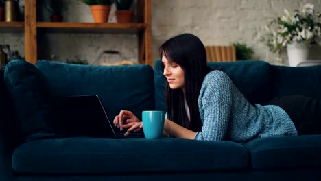 Attractive-young-woman-in-casual-clothes-is-typing-on-laptop-and-smiling-while-lying-on-sofa-in-loft-style-studio.-Modern-technology-and-millennials-concept