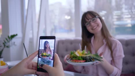 dieta-saludable-y-los-blogs,-chica-alegre-en-gafas-posando-con-frutas-frescas-en-placa-para-novia-que-toma-fotos-en-el-teléfono-móvil-para-redes-sociales-en-café