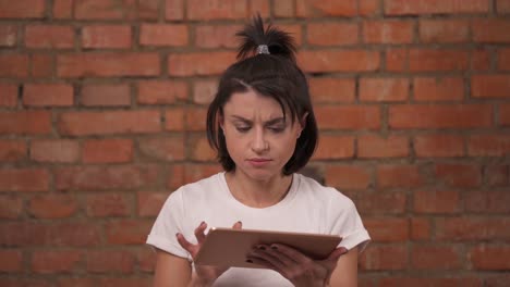 A-young-girl-in-shock-while-using-tablet-device-standing-isolated-against-a-brick-wall-background.