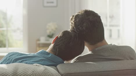 Hinteren-Schuss-süße-männliche-Queer-paar-auf-einem-Sofa-sitzt-und-stecken-ihre-Köpfe-zusammen.-Sie-sind-lässig-gekleidet-in-Jeans-und-Pullover.-Zimmer-ist-Modern-eingerichtet-und-es-ist-hell-und-sonnig.