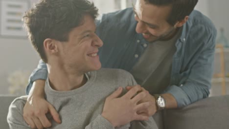 Sweet-Male-Queer-Couple-Spend-Time-at-Home.-Young-Man-Uses-a-Laptop,-His-Partner-Comes-From-Behind-and-Gently-Embraces-Him.-They-Laugh-and-Touch-Hands.-Room-Has-Modern-Interior.