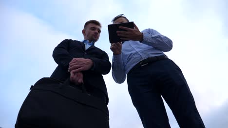 Young-businessman-showing-a-presentation-on-screen-of-tablet-pc-to-his-colleague-in-city.-Successful-entrepreneurs-working-on-digital-tablet-with-blue-sky-at-background.-Low-angle-view-Slow-motion