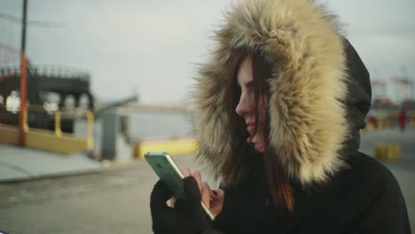 Close-up-shot-of-beautiful-woman-using-smart-phone-technology-app-walking-on-the-street-in-a-warm-jacket-with-a-fluffy-hood