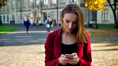 Junge-Studentin-steht-Messaging-am-Telefon,-wird-irritiert-und-lächelt-dann,-außerhalb-der-Universität-an-einem-sonnigen-Tag