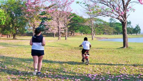 An-Asian-woman-jogging-in-natural-sunlight-in-the-evening,-along-with-his-son-riding-a-bicycle.--exercising-for-good-health.-Slow-Motion