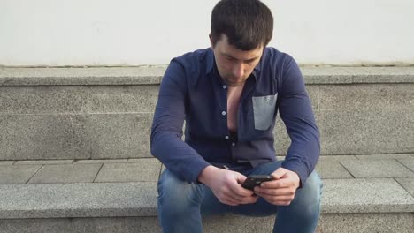 theme-is-man-and-social-networks.-young-Caucasian-sad-male-brunette-in-shirt-is-sitting-in-park-in-Letnec-waiting,-being-late-is-using-hand-to-hold-mobile-phone.-Emotion-resentment-and-disorder