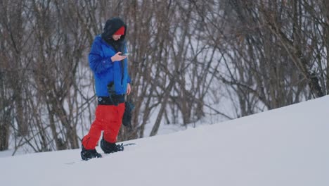 El-snowboarder-masculino-monta-el-tablero-en-el-esquí-hasta-la-ladera-de-la-nieve-y-escribe-mensajes-al-smartphone-a-tus-amigos