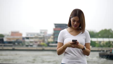 Junge-asiatische-Frau,-die-mit-Smartphone-Spaß-am-Lesen-von-sozialen-Medien-am-Zaun-am-Fluss.