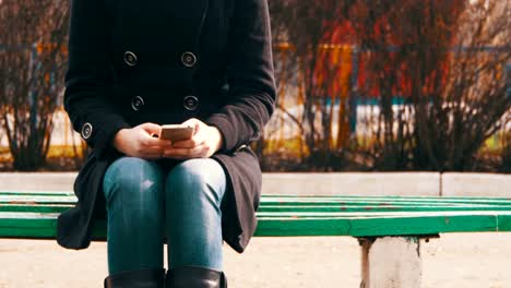 Chica-usando-un-teléfono-móvil-en-un-banco-en-el-parque-de-la-ciudad-en-la-primavera