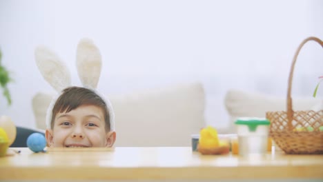 Young-adorable-boy-is-hiding-under-the-table-full-of-Easter-decorations-and-is-playing-with-Easter-bunny-in-his-hand.-Bunny-theatre.