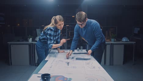 In-der-Dark-Industrial-Design-Engineering-Facility-Male-und-weiblichen-Ingenieurinnen-sprechen-und-arbeiten-Sie-an-einem-Blueprints-Using-Conference-Table.-Auf-dem-Desktop-Zeichnungen,-Entwürfe-und-Elektromotoren-Komponenten,-Teile