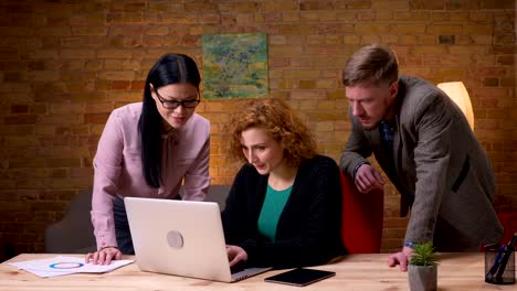 Tiro-de-primer-plano-de-la-joven-empresaria-trabajando-en-la-Laptop-consiguiendo-feliz-y-celebrando-el-éxito-con-dos-colegas.-Empleada-femenina-sosteniendo-una-tableta