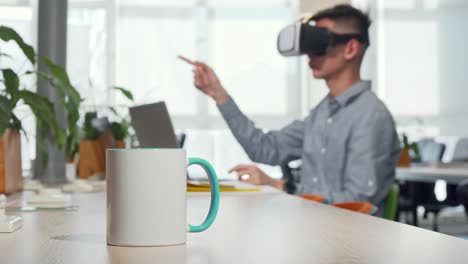 Taza-de-café-en-la-mesa,-hombre-usando-vasos-3d-vr-en-el-trabajo-en-el-fondo