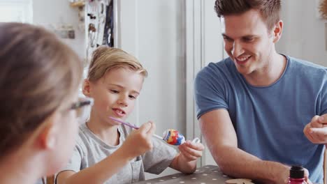 Family-sitting-around-kitchen-table-with-parents-helping-children-to-paint-eggs-for-Easter---shot-in-slow-motion