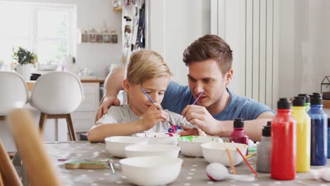 Father-sitting-at-kitchen-table-helping-son-to-paint-eggs-for-Easter---shot-in-slow-motion