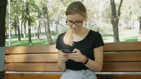 Mujer-en-gafas-chateando-en-el-teléfono-móvil.Relaxed-chica-sentarse-y-mirando-el-teléfono-móvil-en-el-parque-de-la-ciudad.-Hermosa-mujer-tener-chat-usando-Smartphone-Outdoors.Woman-charlando-con-un-amigo.