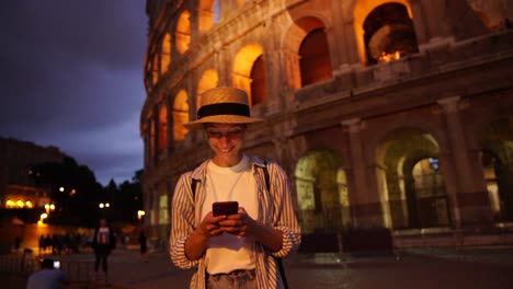 Cheerful-hipster-girl-traveler-enjoying-trip-share-media-in-her-blog-via-smartphone-connected-to-4G-in-roaming-walking-near-coliseumю