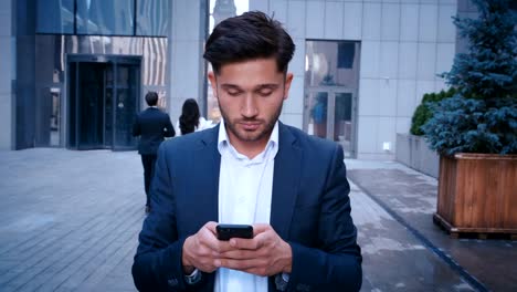 Young-Business-Man-Uses-Smartphone-While-Walking-on-the-Big-City-Business-District-Street.-Classically-Dressed.-Big-Office-Building-in-the-Background.-Looking-Successful,-Confident.