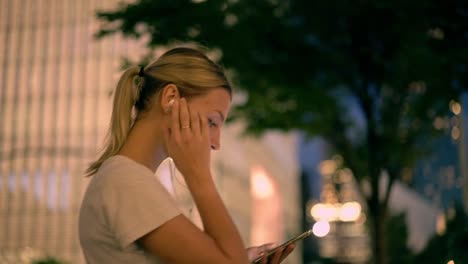 Positive-hipster-girl-listening-favourite-music-via-electronic-headphones-during-strolling-around-evening-city