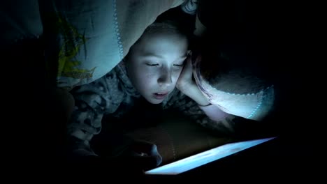 A-child,-a-little-girl,-reads-a-fairy-tale-on-a-tablet-at-night-under-a-blanket-on-the-bed.-Concept-video.-Close-up.-Raw-video.-4K.