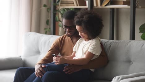 Glückliches-Kind-Mädchen-halten-Telefon-sitzen-auf-afrikanischen-Vater-Schoß