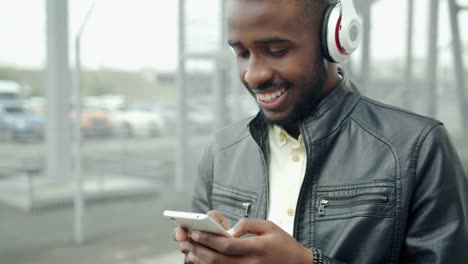 Guapo-chico-en-auriculares-usando-teléfono-inteligente-riendo-en-la-calle-de-la-ciudad