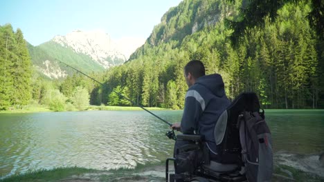 4k-Auflösung-von-behinderten-Fischer-in-einem-elektrischen-Rollstuhl-Angeln-in-schönen-See-in-der-Nähe-von-Wald-und-Berg-im-Rücken