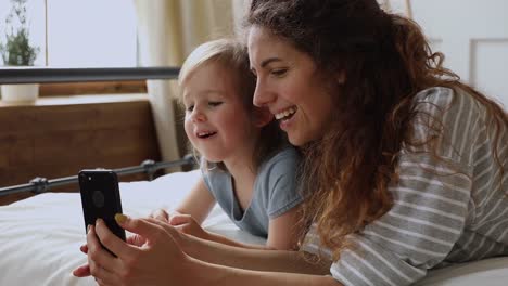 Feliz-mamá-con-hija-de-niño-disfrutar-de-usar-el-teléfono-en-la-cama