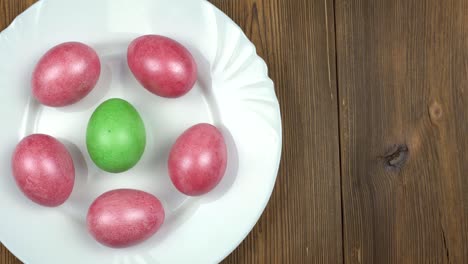 Male-hand-puts-on-the-plate-a-green-Easter-egg-on-the-background-of-red-mother-of-pearl-eggs-in-a-plate,-wooden-background,-copy-space