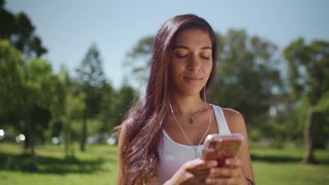 Portrait-of-a-happy-brunette-young-woman-standing-in-the-park-with-earphones-in-her-ears-texting-message-on-smartphone---young-college-student-on-her-phone