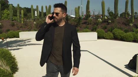 Businessman-walking-in-street-on-holiday-sunny-summer,-black-suit-and-green-shirt,-black-sunglasses.-Using-smartphone-and-having-phone-conversation.-Follow-up-4K-UHD.-Medium-Shot.