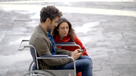 Hombre-sonriente-en-silla-de-ruedas-usando-smartphone-con-su-amigo-en-la-calle