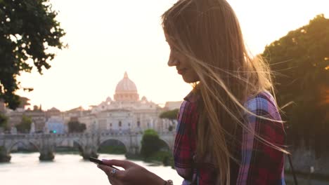 Junge-blonde-Touristin-sitzt-mit-ihrem-Smartphone-auf-einer-Brücke-am-FlussTiber-in-Rom.-Blick-auf-St.-Peter-und-Vatikanstadt,-Italien.