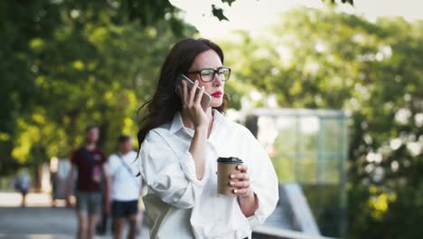 Chica-con-gafas-y-camisa-blanca-hablando-en-el-teléfono-inteligente,-sosteniendo-una-taza-de-café,-teniendo-una-rotura-en-el-parque-con-árboles-verdes.-Concepto-de-negocio.-Cámara-lenta