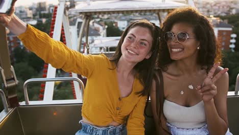 Females-smiling,-showing-victory-sign,-sending-an-air-kiss-while-taking-selfie-on-cell-phone.-Sitting-in-a-ferris-wheel-booth.-Close-up,-slow-motion