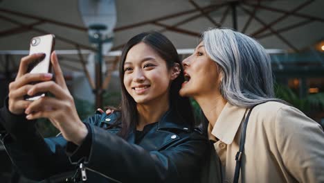 Asian-middle-aged-mother-and-adult-daughter-taking-selfie-by-smartphone-and-smiling-while-posing-near-terrace-of-cafe