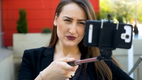 Vista-de-cerca-de-chica-posa-y-hace-muecas-con-smartphone-cámara-y-selfie-palo-en-la-ciudad