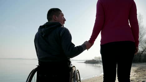 Disabled-man-in-wheelchair-gives-hand-to-girlfriend,-back-view,-near-river