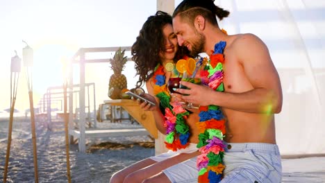 mobile-communication,-girl-sitting-with-guy-on-beach-and-holding-in-hands-cell-phone,-beautiful-couple,-young-people-from-Hawaii-lei-on-neck-drink-cocktail-View-social-networks