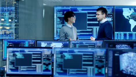 In-the-System-Control-Room-Project-Manage-and-IT-Engineer-Have-Discussion,-they're-surrounded-by-Multiple-Monitors-with-Graphics.-They-Work-in-a-Data-Center-on-Data-Mining,-AI-and-Neural-Networking.